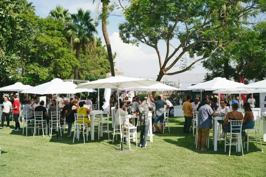 People gather on the grass around white high stoor and bar tables and white umbrellas at an outdoor event