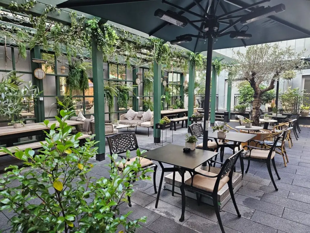 An outdoor quiet space. A covered courtyard with sofas and bistro tables. There are plants surrounding the tables and on the back wall.